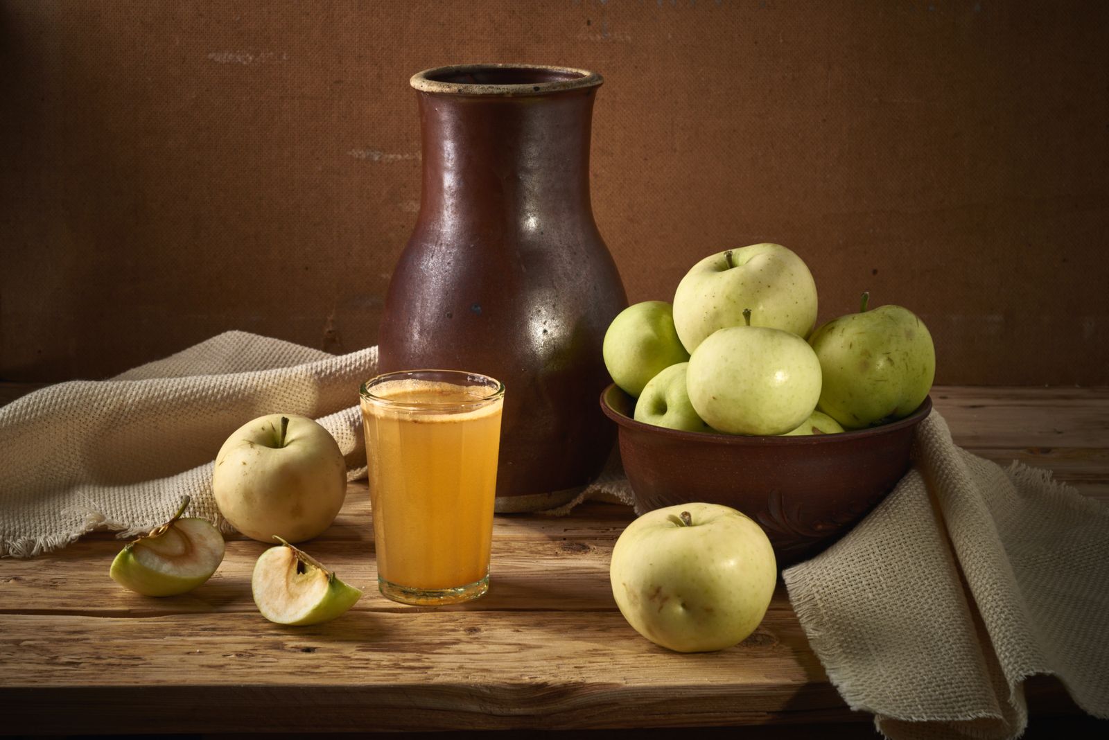 Glas naturtrüber Apfelsaft neben Tonkrug und Schale mit grünen Äpfeln auf einem Holztisch