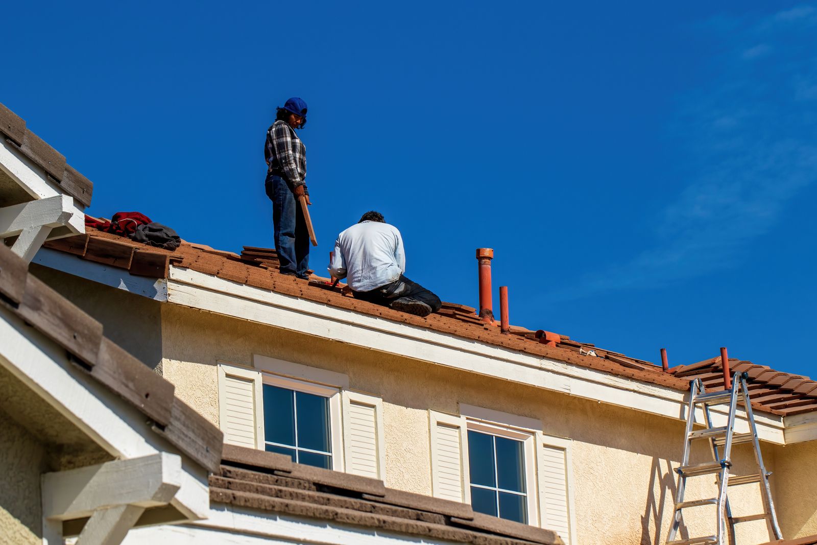 Zwei Dachdecker arbeiten unter blauem Himmel auf einem Ziegeldach über der Fassade eines Wohnhauses