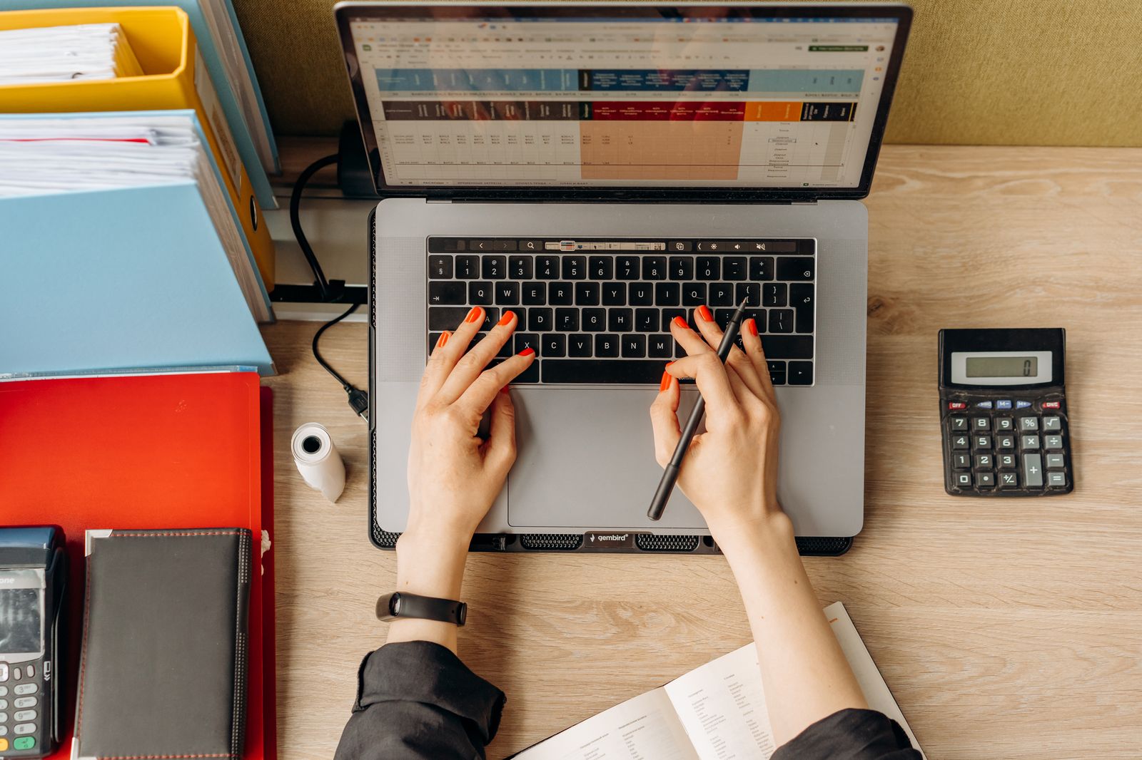 Hände tippen auf einer Laptop-Tastatur, daneben Aktenordner und ein Taschenrechner auf dem Schreibtisch