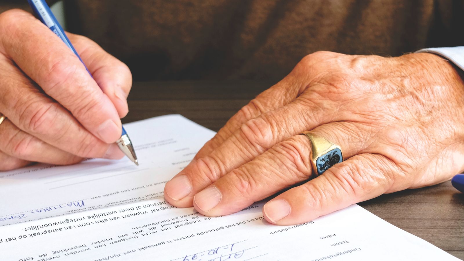 Ältere Hand mit goldenem Ring unterschreibt ein Dokument am Schreibtisch