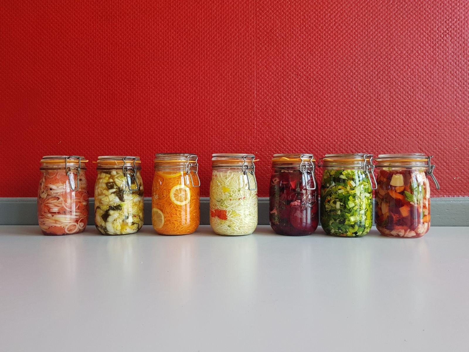 Bunte Einmachgläser mit fermentiertem Gemüse nebeneinander vor roter Wand