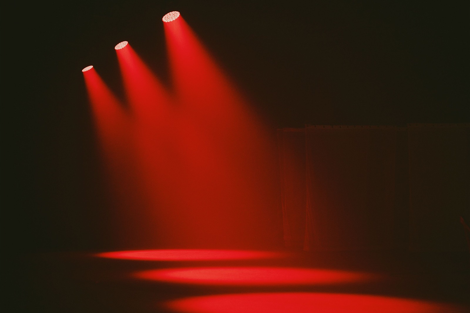 Drei rote Scheinwerfer werfen Lichtkegel auf eine dunkle, leere Theaterbühne
