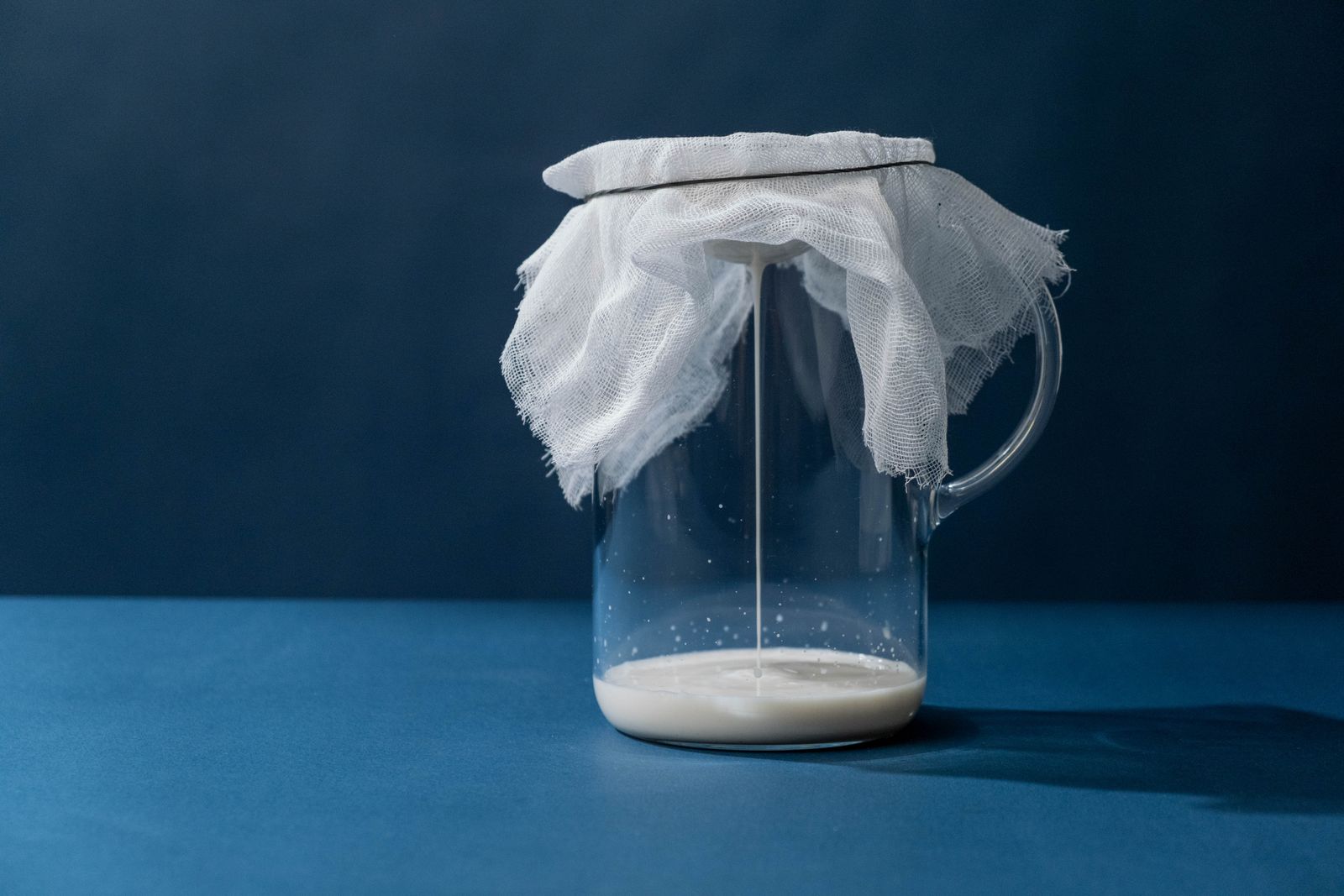 Milch wird durch ein Käsetuch in einen Glaskrug gefiltert, daneben eine Schale mit Molke