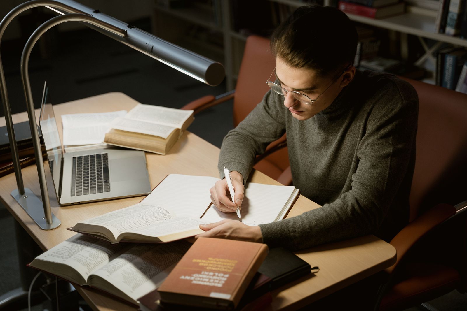 Junger Mann schreibt Notizen in ein Heft beim Lesen eines Buches am Schreibtisch