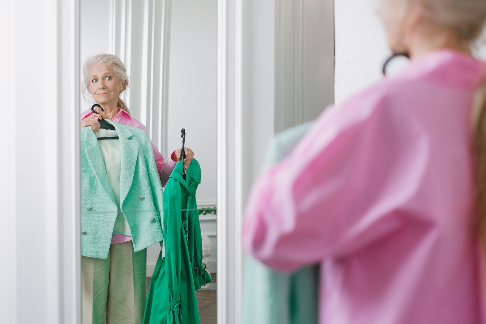 Ältere Frau mit grauem Haar hält vor dem Spiegel einen grünen Blazer und wählt ihr Outfit