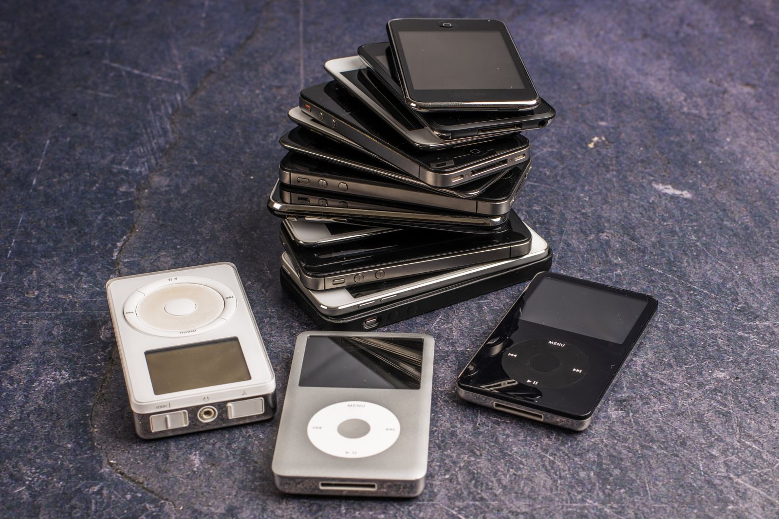 Stapel gebrauchter Smartphones neben drei älteren MP3-Playern auf dunklem Untergrund