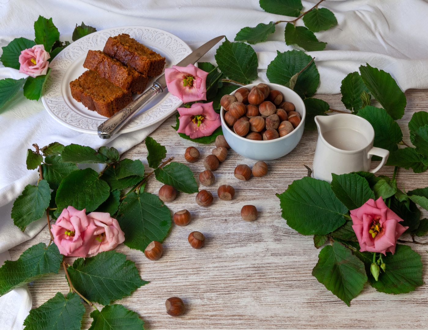 Scheiben Nusskuchen aus der Kastenform auf weißem Teller, daneben eine Schale Haselnüsse und ein Milchkännchen