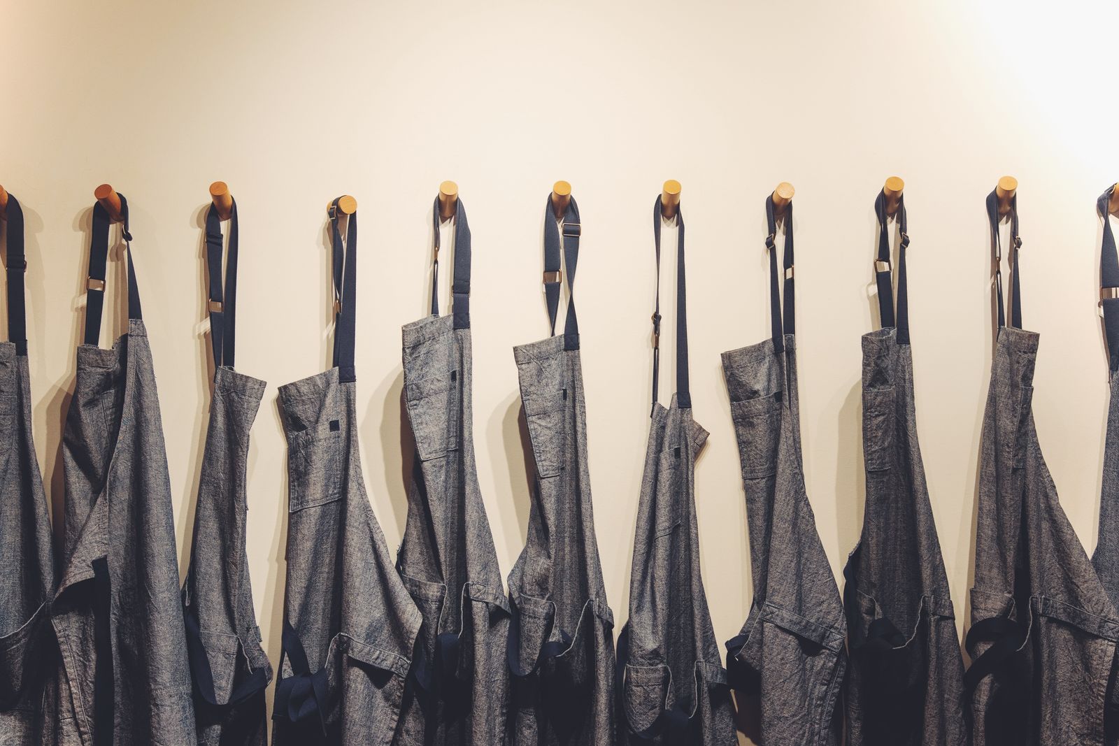 Reihe von Schürzen aus dunklem Denim an Wandhaken vor einer hellen Wand
