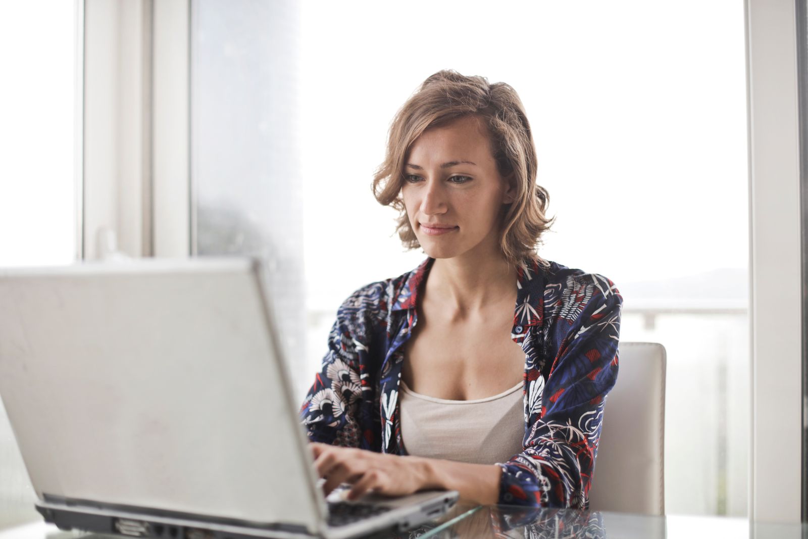 Frau arbeitet an einem Laptop an einem Glastisch vor einem hellen Fenster