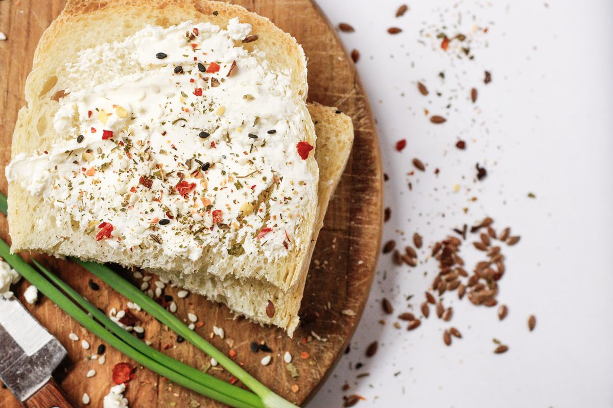 Scheibe Weißbrot mit Frischkäse, bestreut mit Gewürzen und Leinsamen, auf einem Holzbrett neben Frühlingszwiebeln