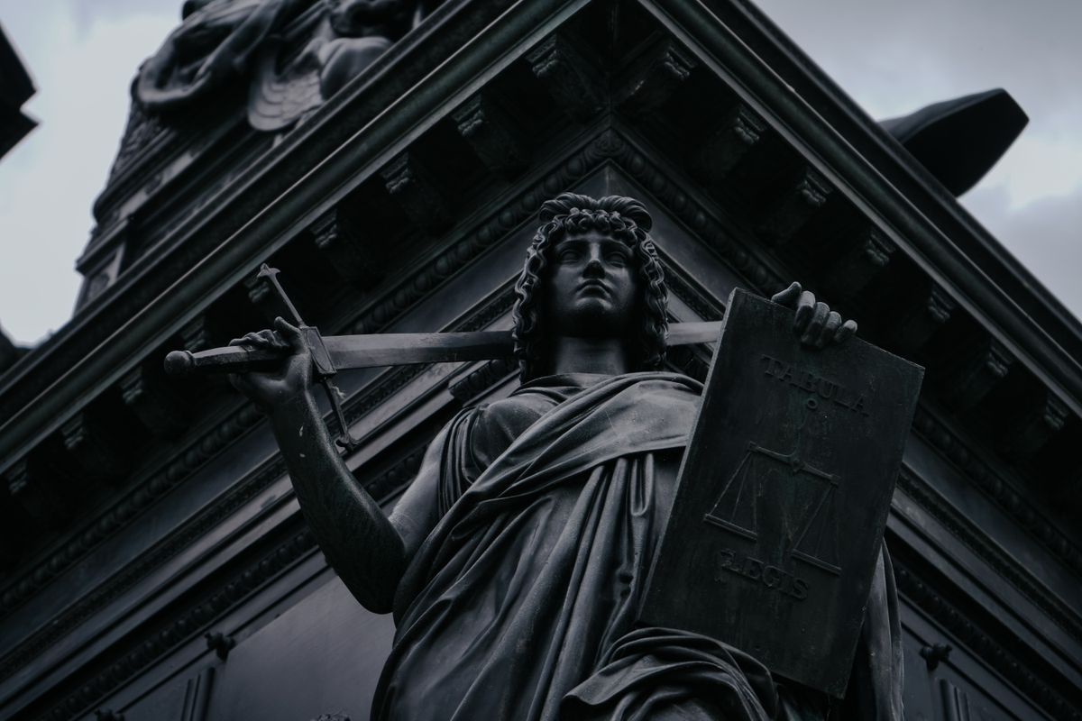 Dunkle Bronzefigur der Justitia mit Schwert und Gesetzestafel vor dem Giebel eines Gerichtsgebäudes, von unten fotografiert