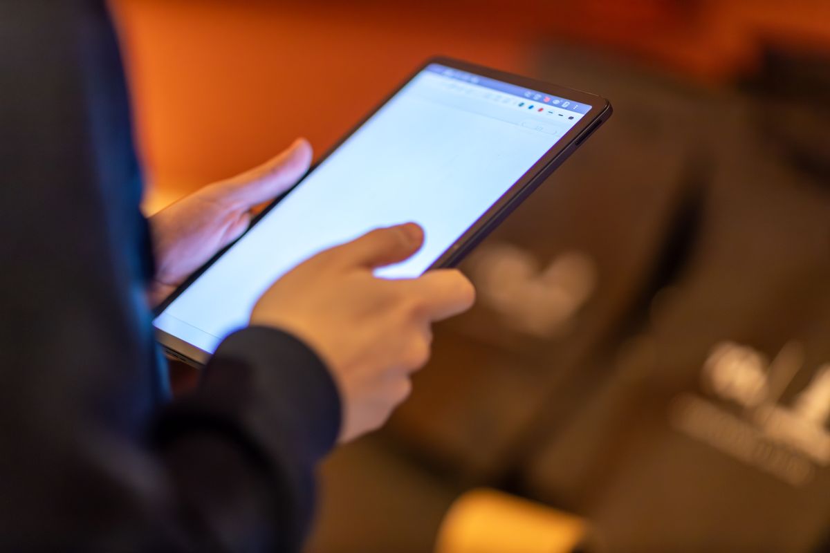 Person hält ein Tablet mit hell leuchtendem Bildschirm in beiden Händen und tippt mit dem Daumen darauf, im Hintergrund unscharfes warmes Licht