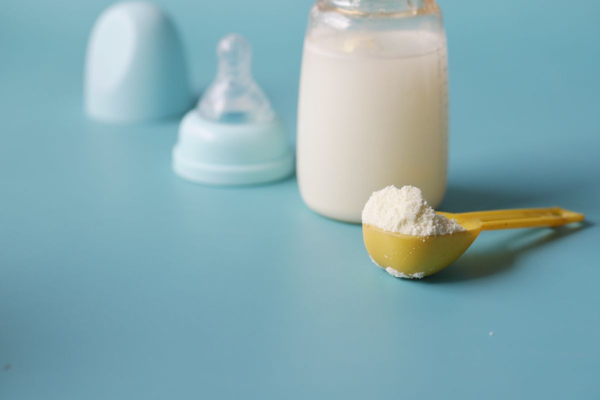 Gelber Messlöffel mit Milchpulver vor einer gefüllten Babyflasche und einem abgenommenen Sauger auf hellblauem Untergrund