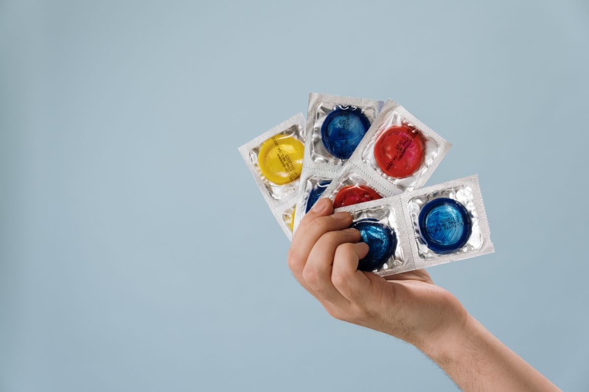 Hand hält mehrere eingeschweißte Kondome mit roten, blauen und gelben Ringen vor einem graublauen Hintergrund