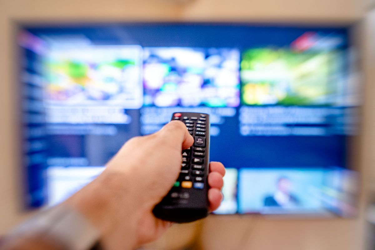Hand richtet eine Fernbedienung auf einen unscharfen Fernseher, auf dem eine Übersicht mit bunten Programmkacheln zu sehen ist