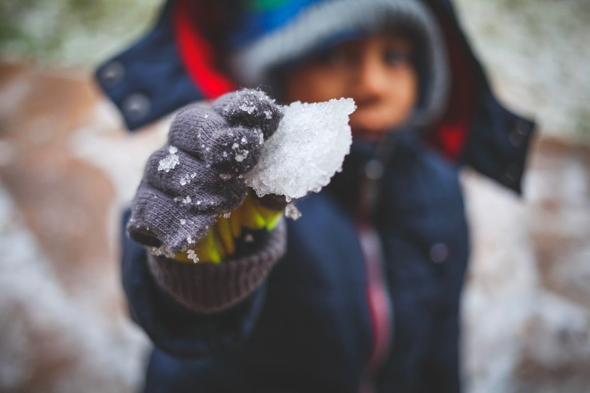 Kind in Winterjacke hält ein Stück Schnee in einer Hand mit grauem Strickhandschuh nach vorn