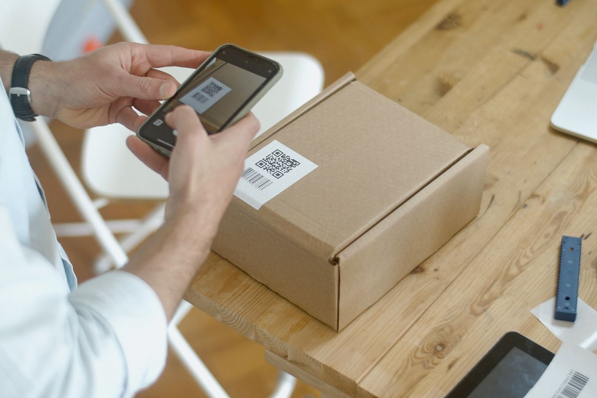 Hand hält ein Smartphone über das Etikett eines Kartons und erfasst den aufgedruckten Code