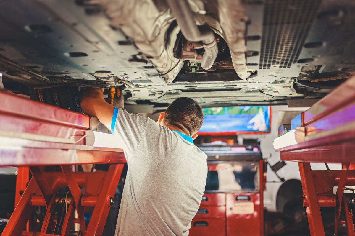 Mechaniker in grauem T-Shirt arbeitet von unten an einem Auto, das auf einer roten Hebebühne steht